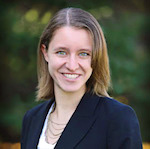 Professional headshot of a woman with shoulder-length brown hair and blue eyes, smiling and wearing a dark blazer over a white top with layered necklaces against a blurred outdoor greenery background.