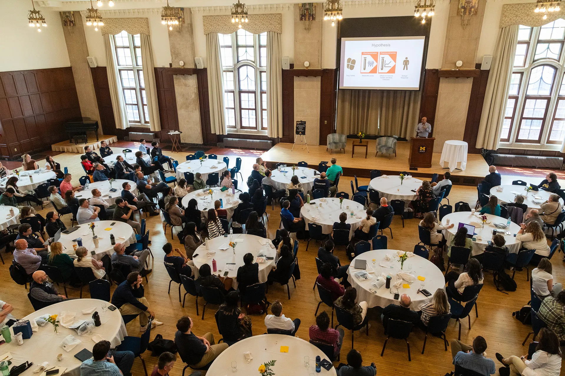 An aerial photo of the Union ballroom filled with attendees sitting at round tables. A presenter stands at a podium on stage.